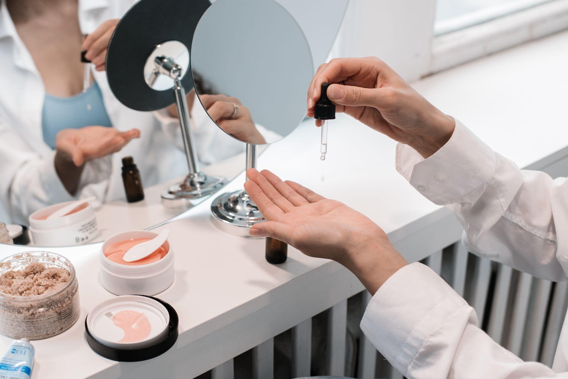Kosmetikerin gibt ein klares Serum aus der Pipette in die Handfläche vor einem Spiegel